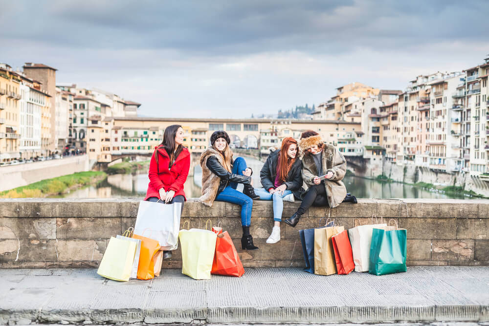 planning a trip to Italy. Four young female friends take a break from shopping for the city shops. Are sitting on the railing of a bridge, two talk while two look phone, shopping bags around them, behind them the Ponte Vecchio
