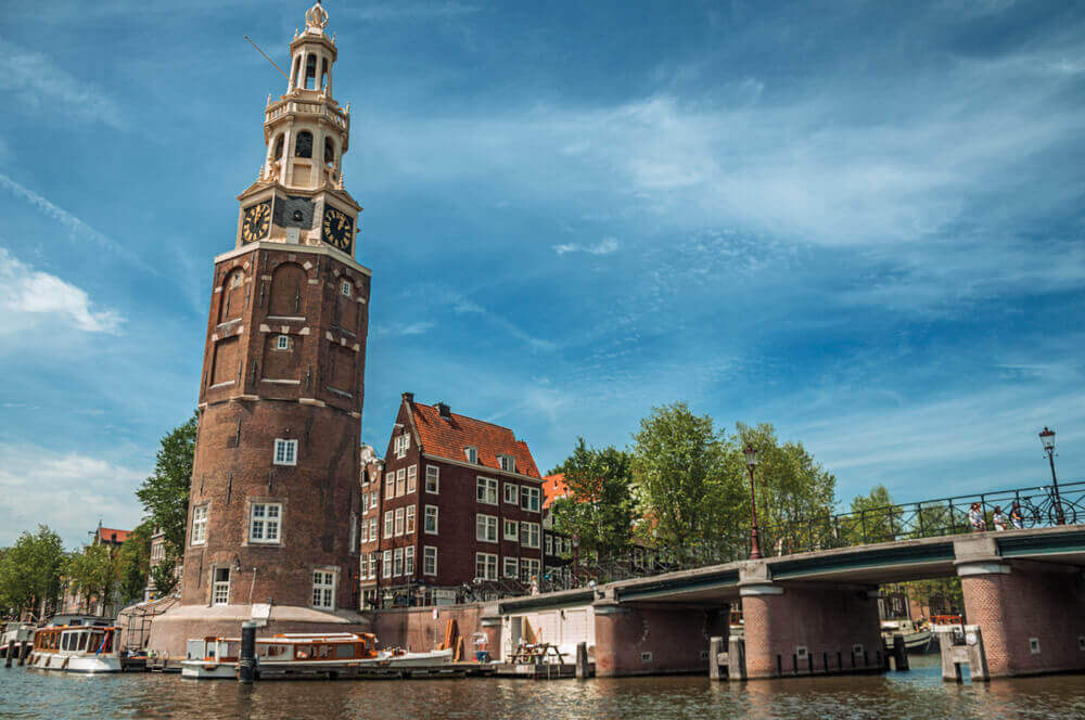 plan a trip to Europe. Old brick bell tower and bridge near the tree-lined canal with moored boats and blue sky in Amsterdam. Famous for its huge cultural activity, graceful canals and bridges. Northern Netherlands.