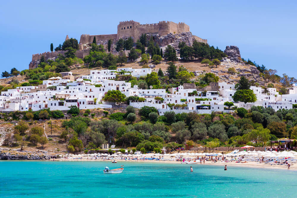 View of the Acropolis Lindos on top a hill, with neighbourhood of low-rise white buildings built into the hill. Sparkling beach in the foreground at the base of the hill. online trip planner.